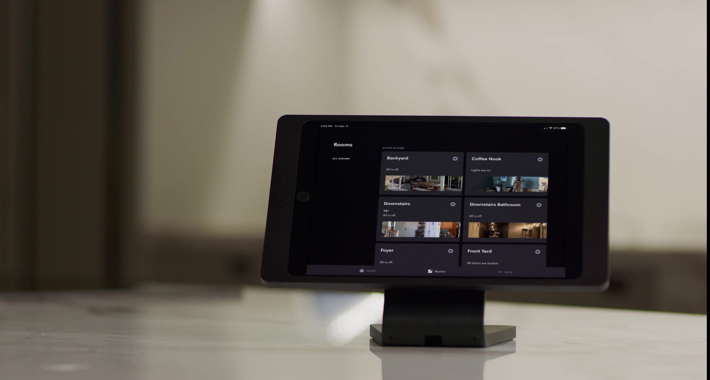 Tablet on a stand displaying a smart-home dashboard with tiles for rooms like Backyard, Coffee Nook, Downstairs, Foyer, and Front Yard—status shows lights and locks—sitting on a marble counter with a softly blurred interior in the background.