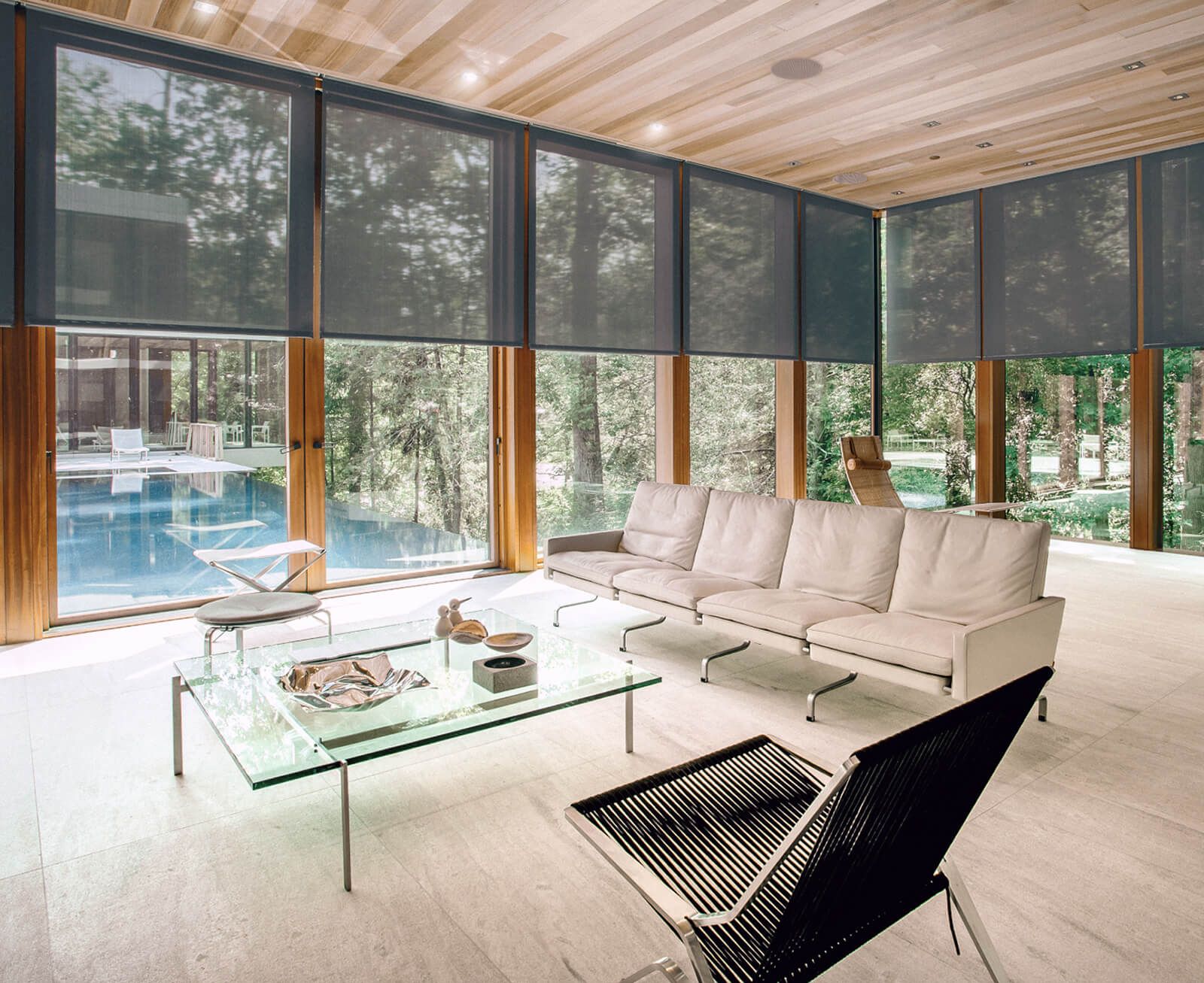 Sunlit modern living room with floor-to-ceiling windows fitted with semi-opaque roller shades, light wood ceiling with recessed lights, a long cream sofa and minimalist glass coffee table; views to a forested yard and a blue swimming pool outside.